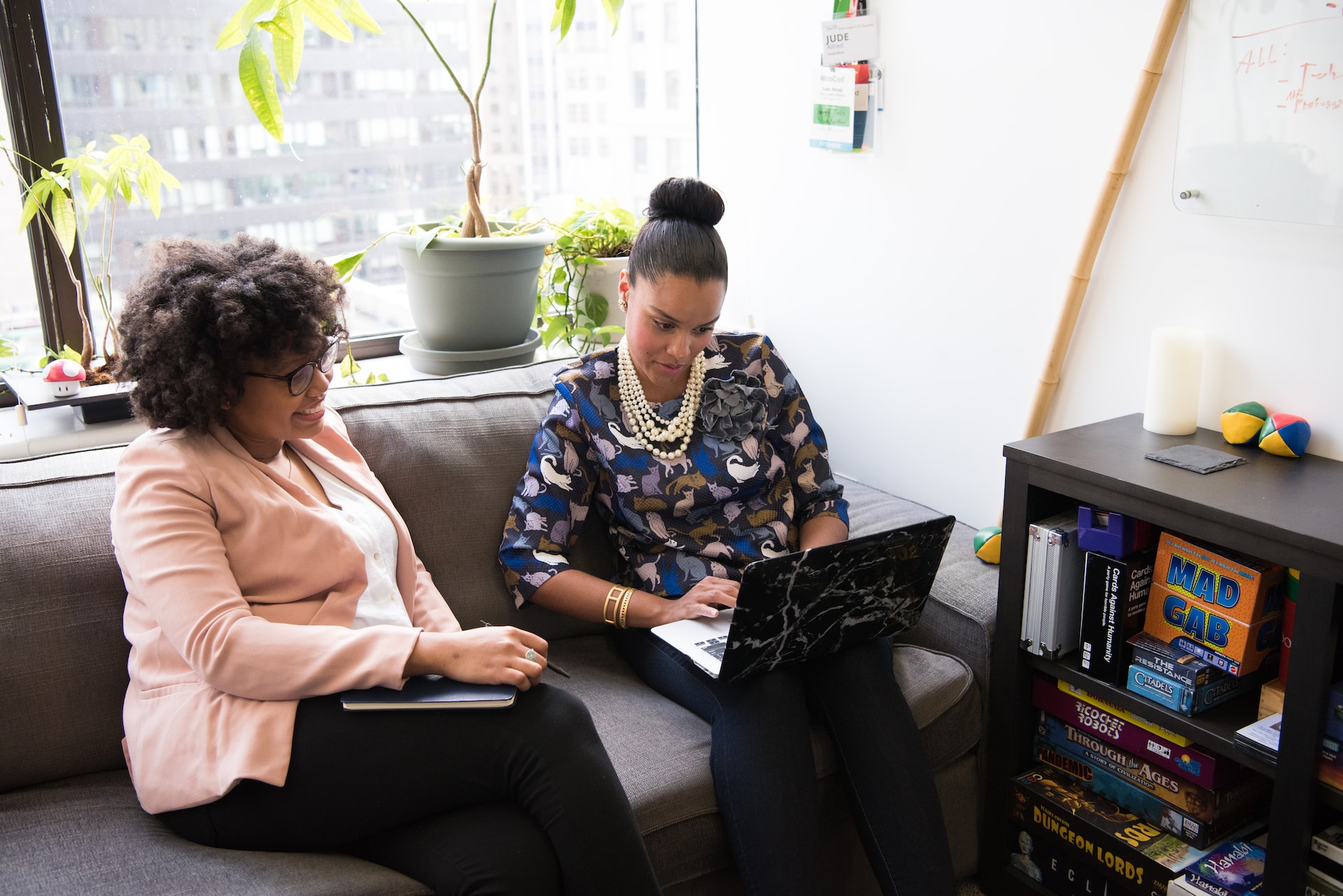 Two professional black women sitting on a couch in an office and looking at a laptop