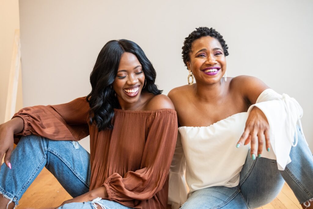 Two confident black women sitting next to eachother, shoulders touching, smiling and laughing