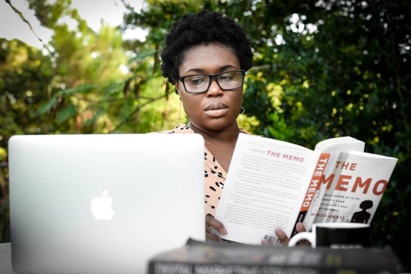 A black woman sitting in front of a computer with a novel in her hand